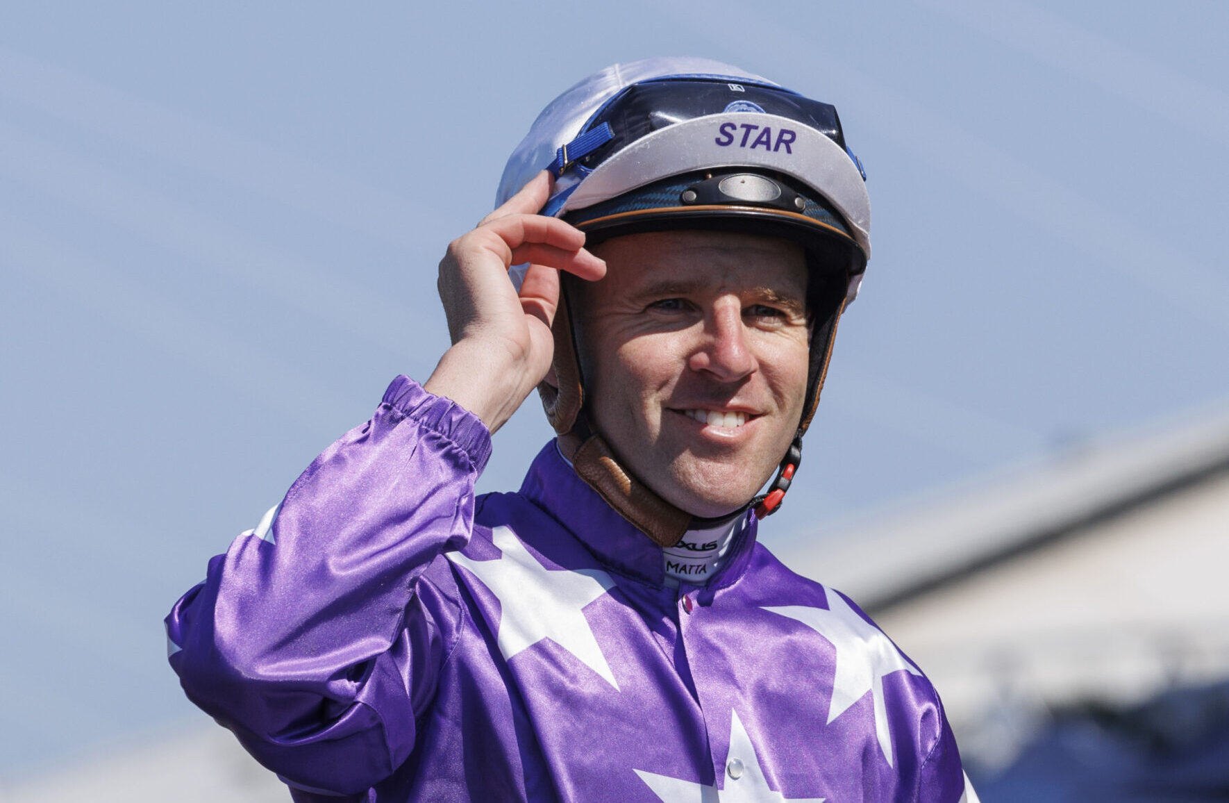 Smiling jockey in purple racing silks adjusts his helmet with STAR on the visor outdoors at a track.