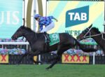 Two jockeys race their horses toward the finish line, one in blue checkered silks on a dark horse wearing a green number 9 saddlecloth, with bright advertising boards behind them.