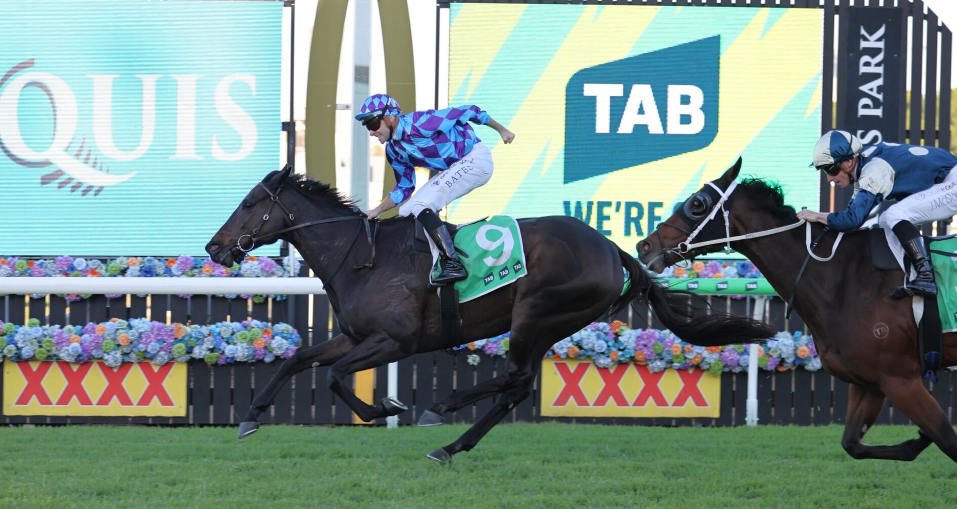 Two jockeys race their horses toward the finish line, one in blue checkered silks on a dark horse wearing a green number 9 saddlecloth, with bright advertising boards behind them.