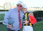 Two winners posing with trophies at a racecourse, smiling with arms around each other in racing attire and caps that say J EWEL RACE DAY (paraphrased).