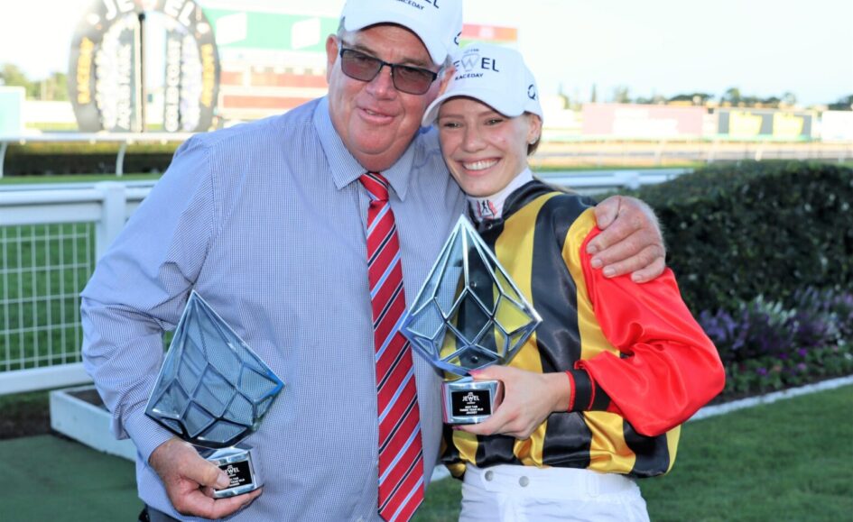 Two winners posing with trophies at a racecourse, smiling with arms around each other in racing attire and caps that say J EWEL RACE DAY (paraphrased).
