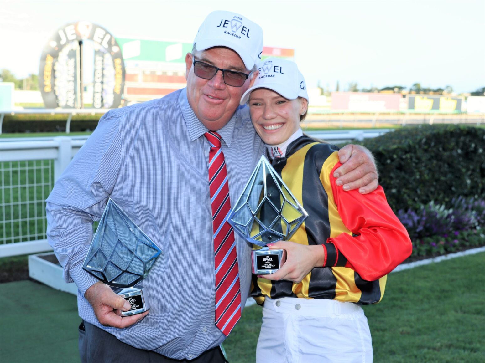 Two winners posing with trophies at a racecourse, smiling with arms around each other in racing attire and caps that say J EWEL RACE DAY (paraphrased).