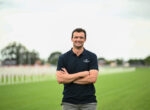 Smiling man in a navy polo with arms crossed on a grassy sports field, stadium/track in the blurred background.