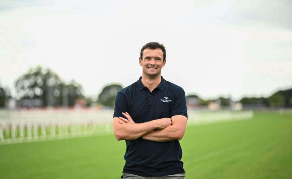 Smiling man in a navy polo with arms crossed on a grassy sports field, stadium/track in the blurred background.