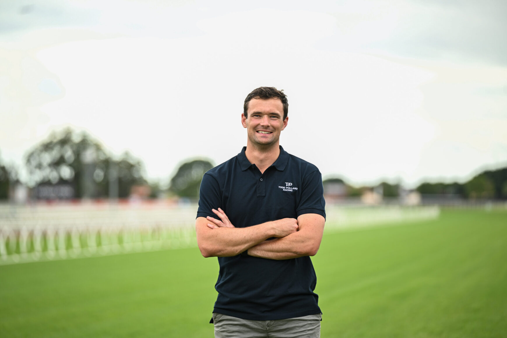Smiling man in a navy polo with arms crossed on a grassy sports field, stadium/track in the blurred background.
