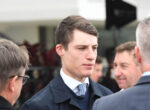 Man in a dark suit and patterned tie speaks with a group at a formal event, foregrounding his focused expression.