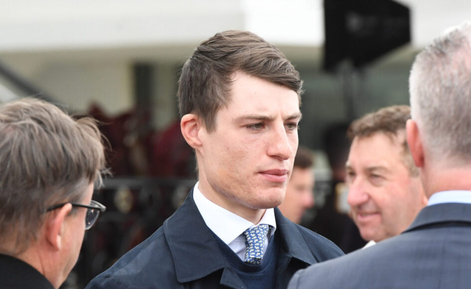 Man in a dark suit and patterned tie speaks with a group at a formal event, foregrounding his focused expression.