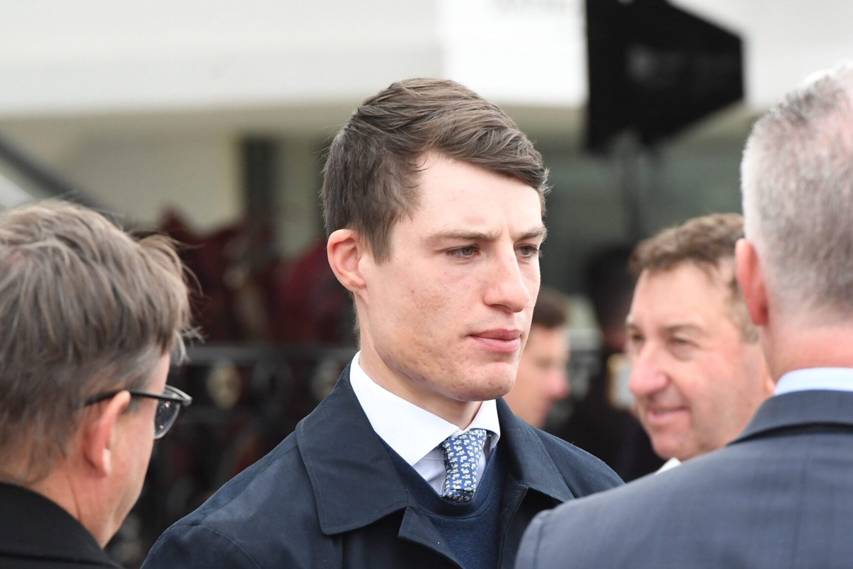Man in a dark suit and patterned tie speaks with a group at a formal event, foregrounding his focused expression.