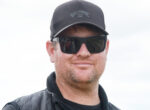 Man wearing a black Billabong cap and dark sunglasses, outdoors, smiling softly.