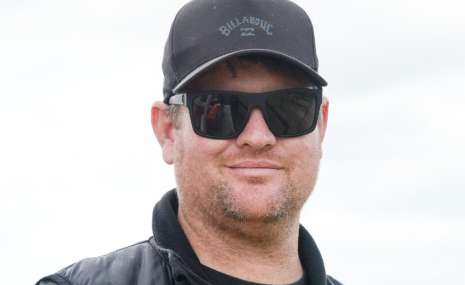 Man wearing a black Billabong cap and dark sunglasses, outdoors, smiling softly.