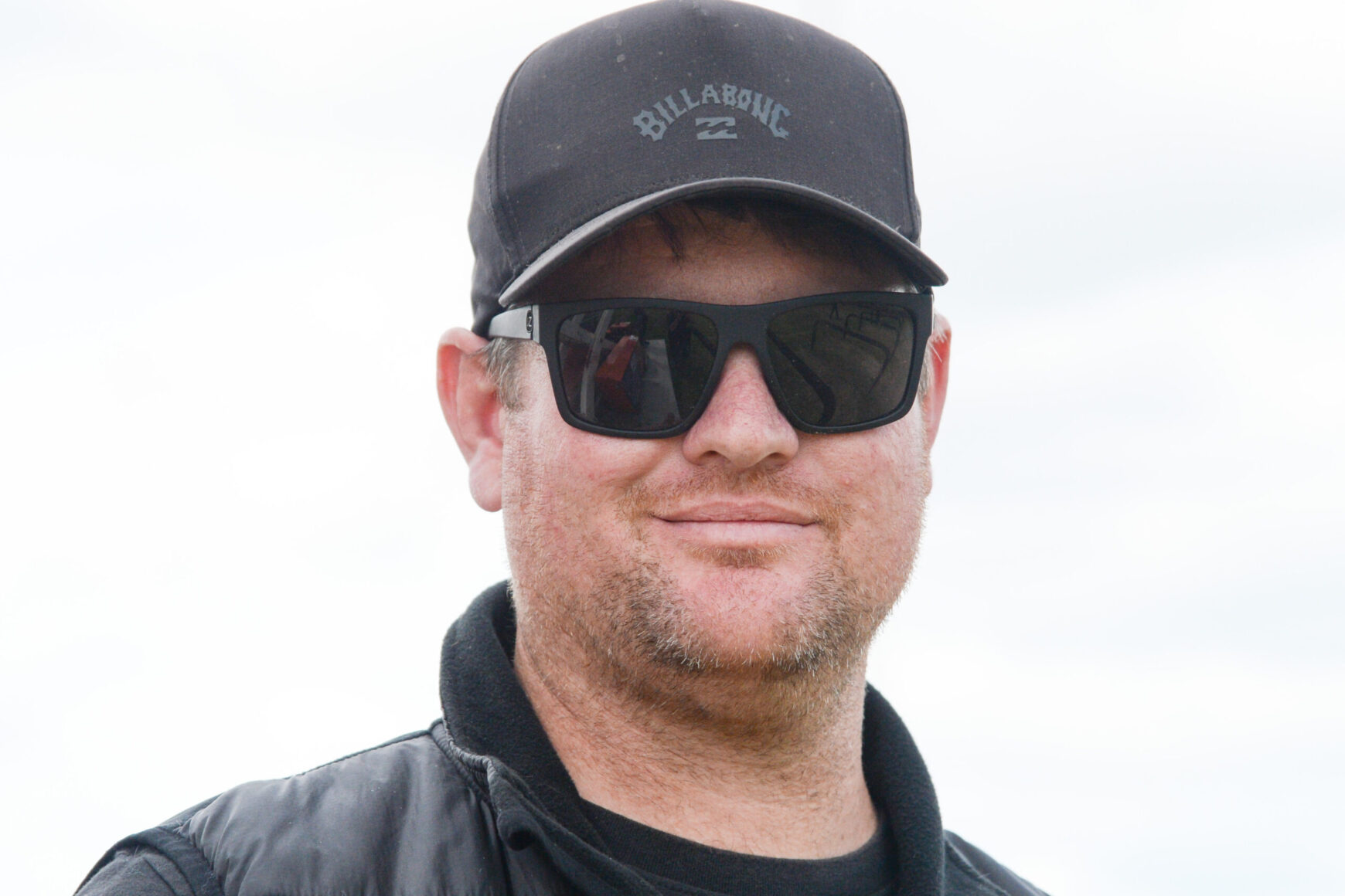Man wearing a black Billabong cap and dark sunglasses, outdoors, smiling softly.