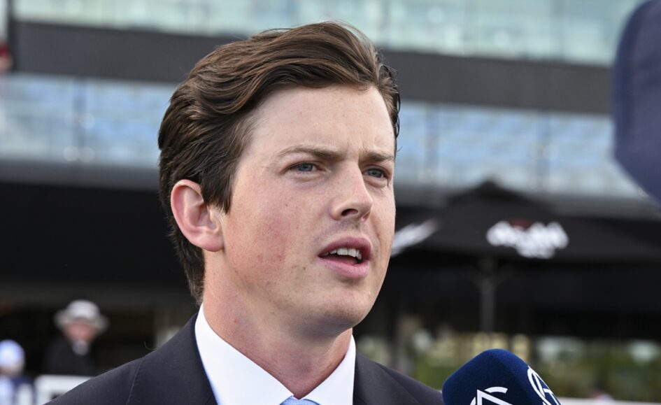 Man in a dark suit and light blue tie speaks into a blue microphone outdoors, with a modern building in the background.",