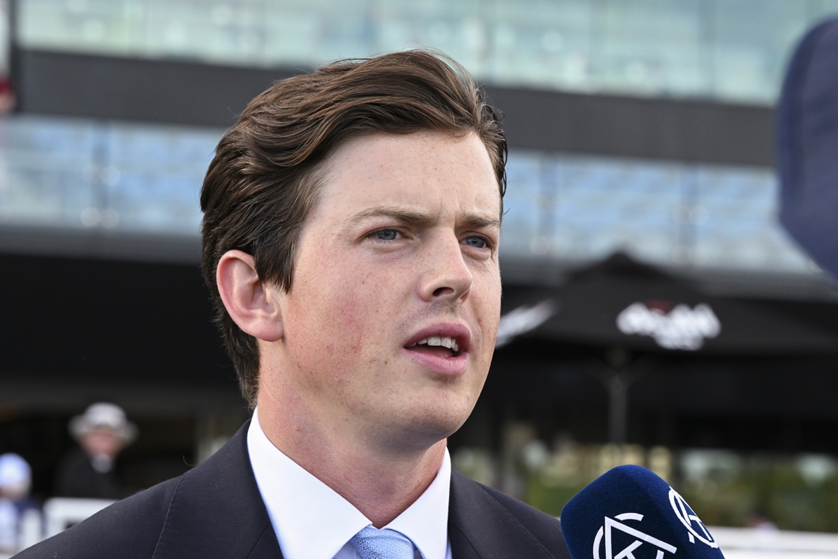 Man in a dark suit and light blue tie speaks into a blue microphone outdoors, with a modern building in the background.",