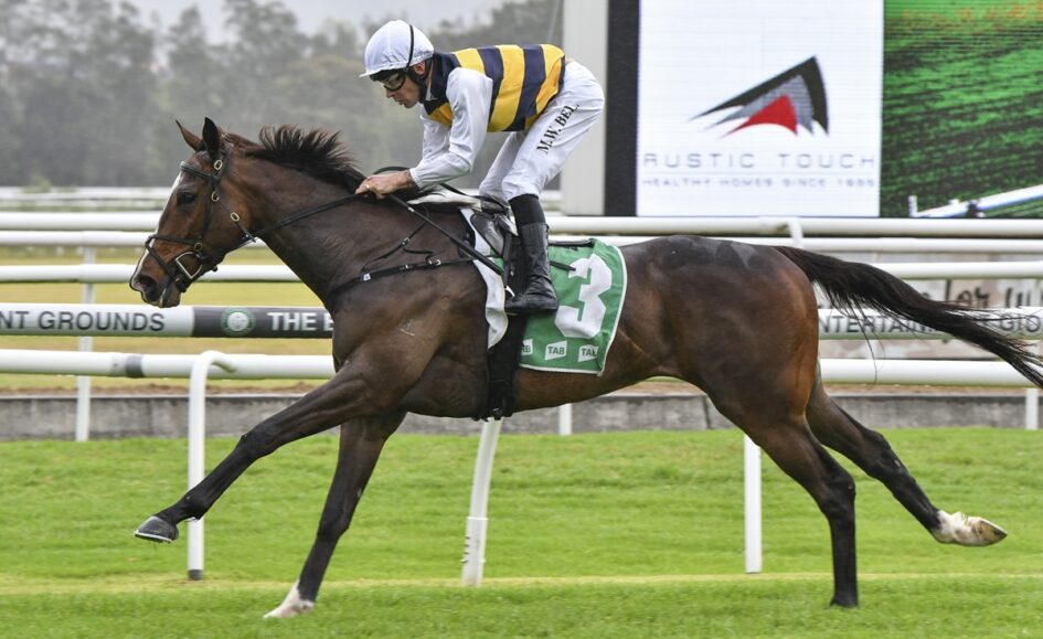 Jockey in white and yellow silks rides a dark brown horse with green number-3 saddlecloth, galloping on a grassy racetrack.