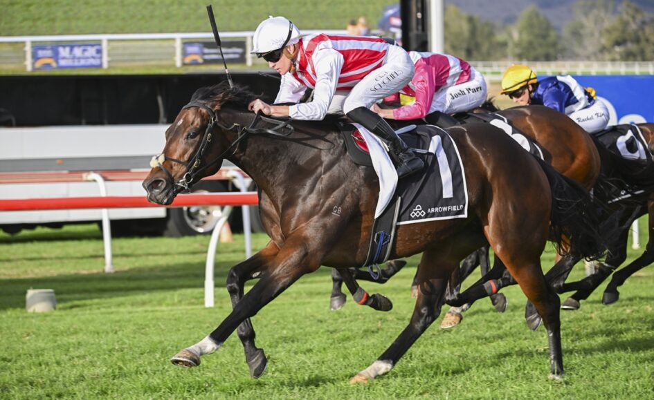 Front-running racehorse with jockey in red-and-white silks sprinting on a grassy track, others close behind on a sunny day.