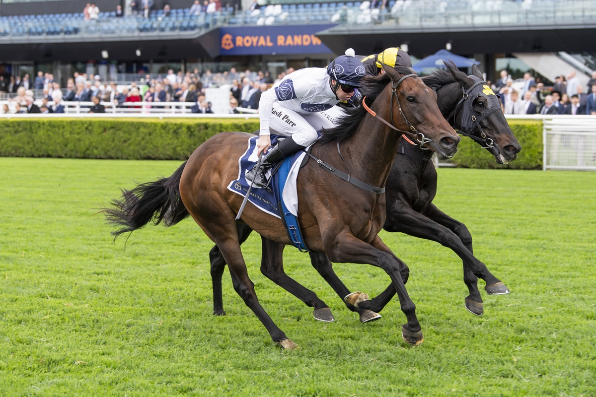 Two racehorses sprint on a green turf as jockeys urge them forward, with a crowd in the stands behind them.