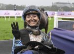 Jockey in a muddy blue riding outfit smiling and flashing a peace sign after a race, helmet and goggles muddy, on a wet racetrack.