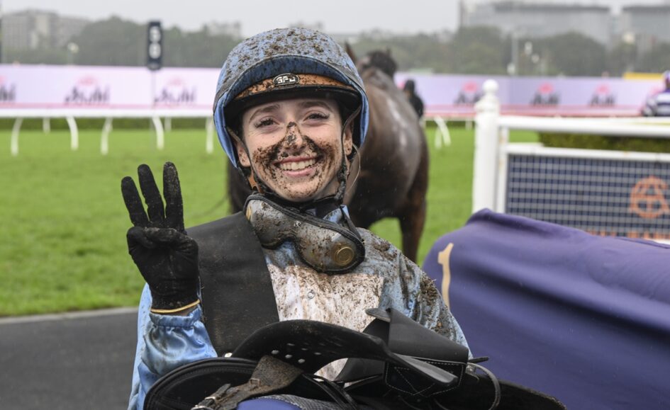 Jockey in a muddy blue riding outfit smiling and flashing a peace sign after a race, helmet and goggles muddy, on a wet racetrack.