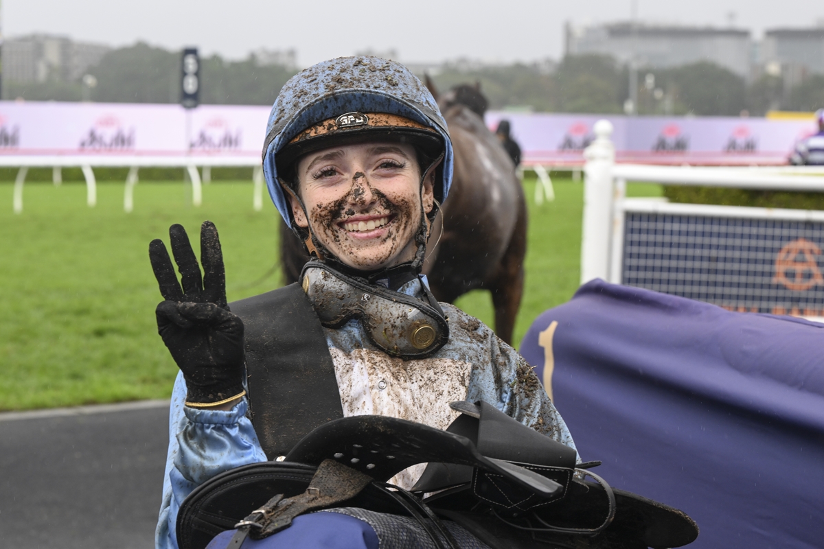 Jockey in a muddy blue riding outfit smiling and flashing a peace sign after a race, helmet and goggles muddy, on a wet racetrack.