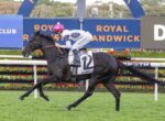 Black horse gallops on a racecourse with jockey in pink cap and white silks, number 12 saddlecloth, at Royal Randwick signage behind.