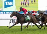 Horse race in progress: lead jockey in red silks on a dark bay horse wearing a green number 8 saddlecloth, chased by rivals on a grassy track.