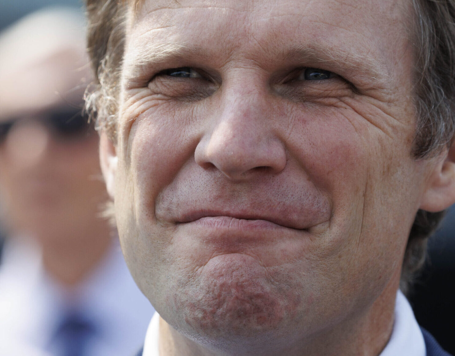 Close-up of a middle-aged man making a puckered lips expression, squinting slightly outdoors, with visible wrinkles and stubble.