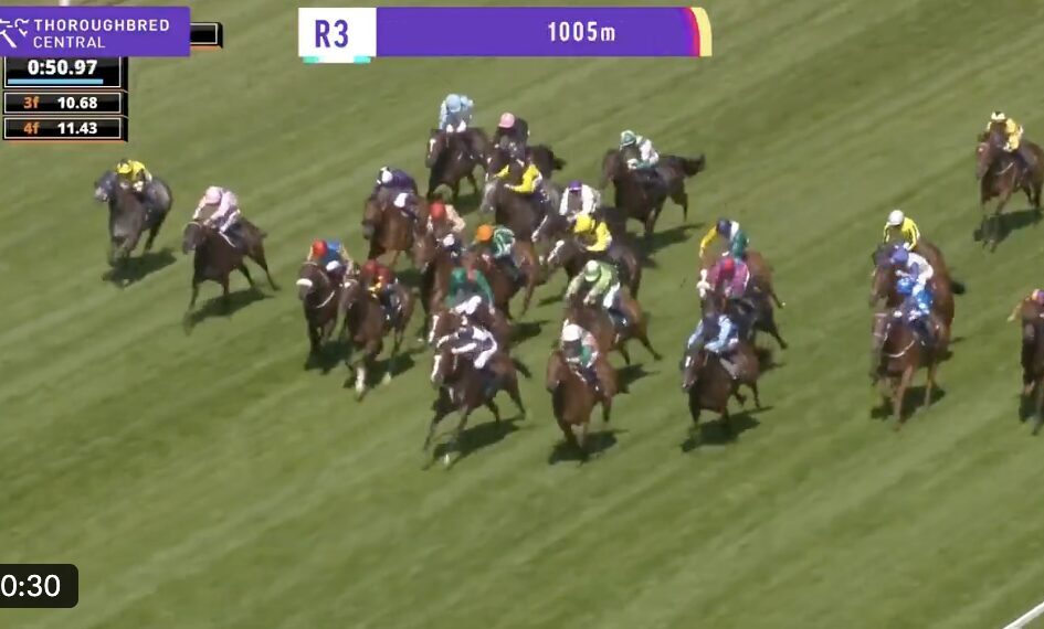 Group of thoroughbred horses racing on a green turf track with jockeys in colorful silks; on-screen graphics show 'R3' and '1005m'