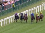 Seven thoroughbred horses race along the turf at Royal Ascot, jockeys in purple, blue, and yellow silks, with spectators beyond the trackRail fence on the left.