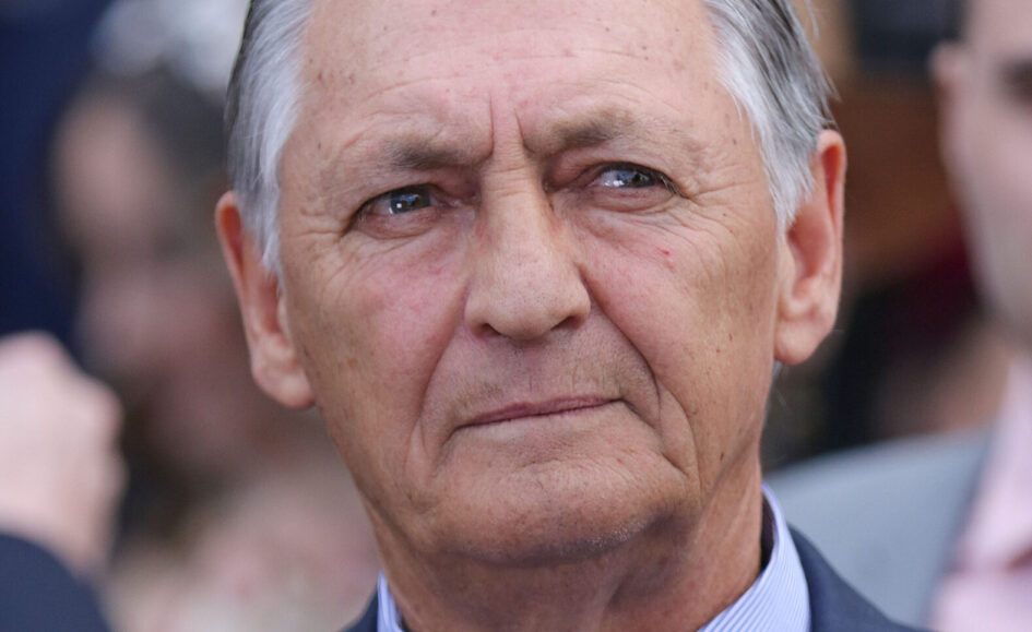 Close-up portrait of an older man with gray hair in a dark suit, looking forward with a thoughtful expression outdoors.