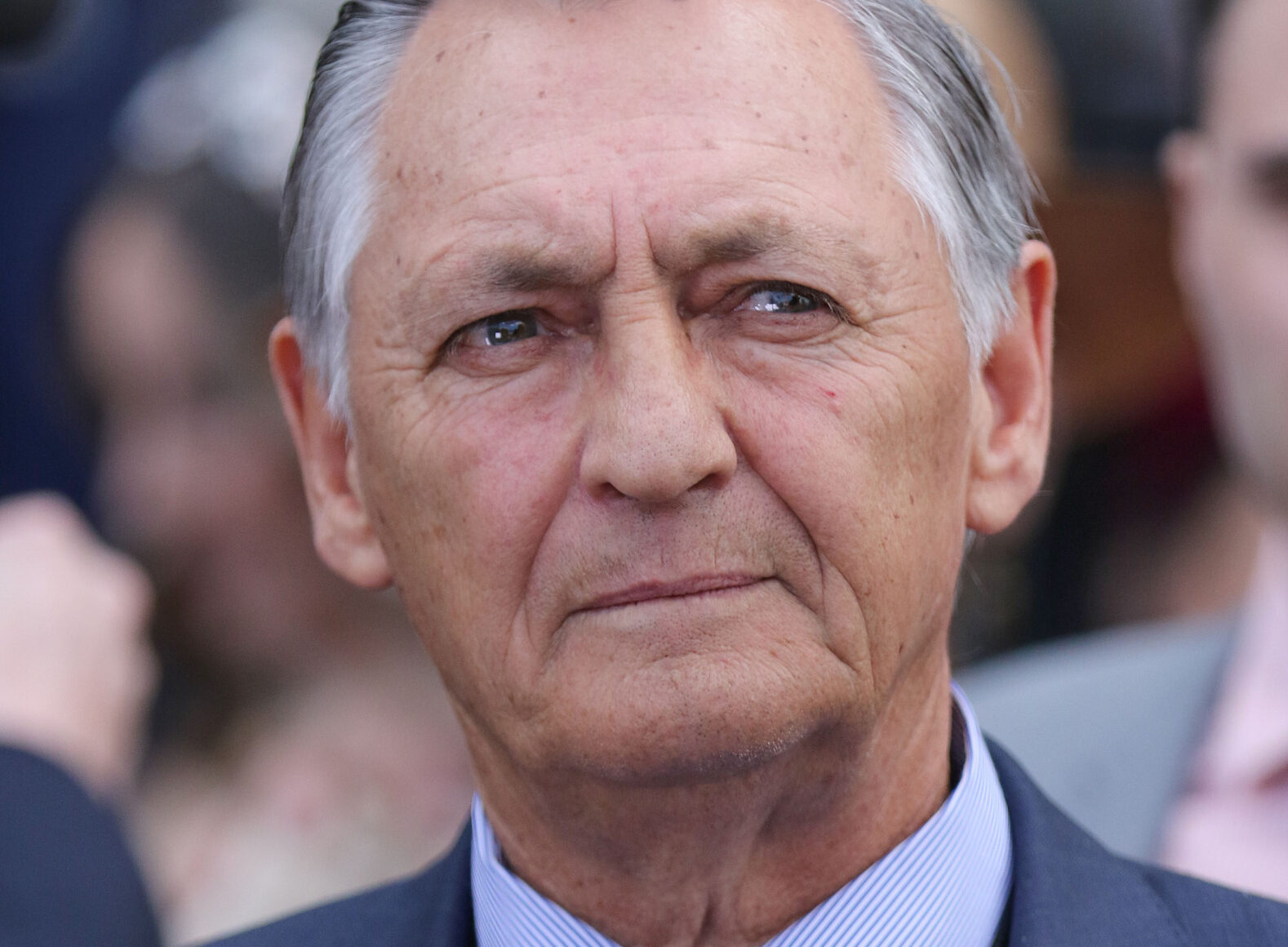 Close-up portrait of an older man with gray hair in a dark suit, looking forward with a thoughtful expression outdoors.