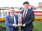 Two men in suits pose outdoors, handing a crystal trophy to celebrate a race win at a sports event.