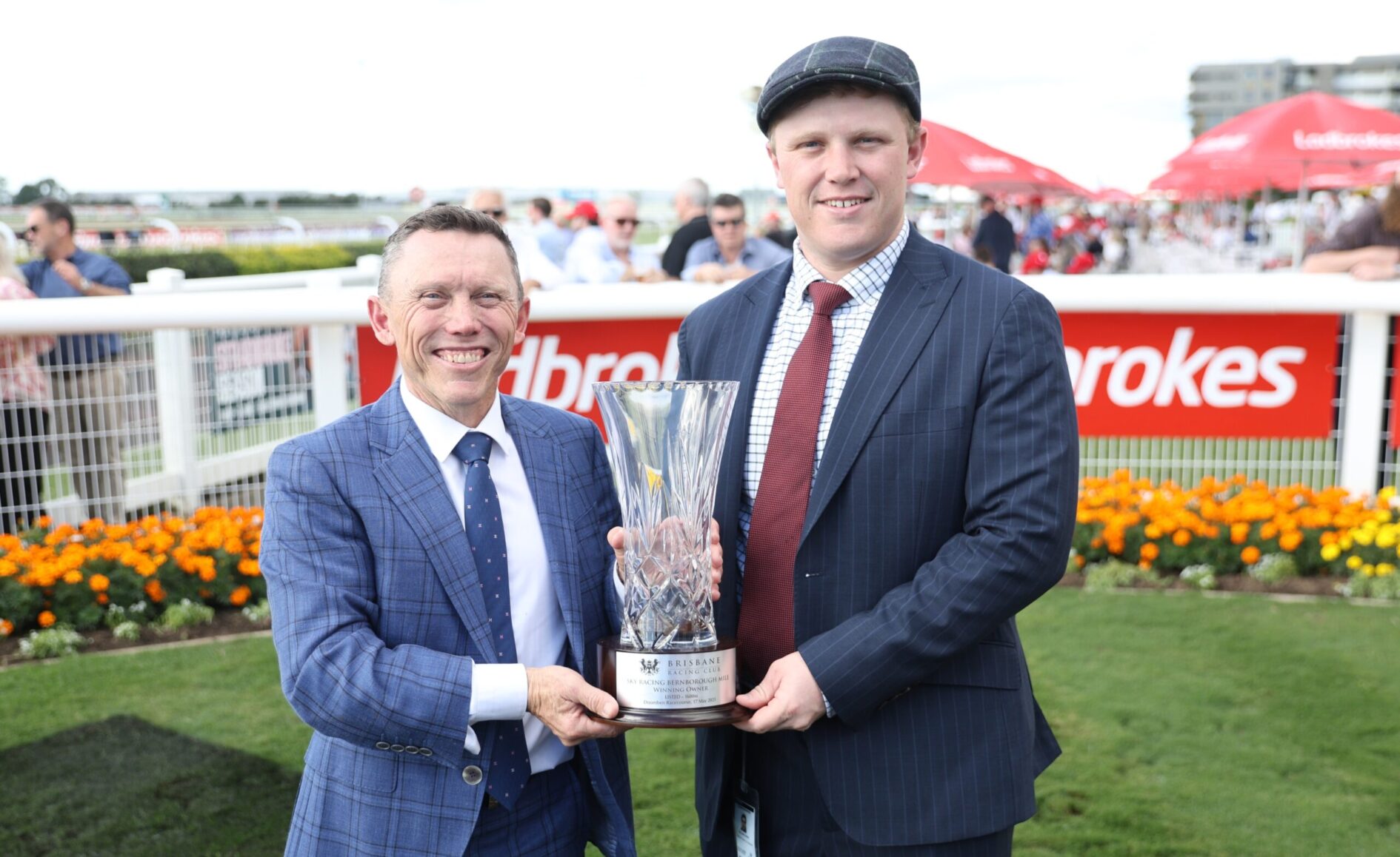 Two men in suits pose outdoors, handing a crystal trophy to celebrate a race win at a sports event.
