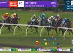 Group of thoroughbred horses racing on a grassy track; broadcast overlay shows race details and horse numbers.