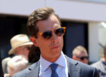 Man in a navy suit and tie wearing orange sunglasses at an outdoor event, with a crowd in the background.