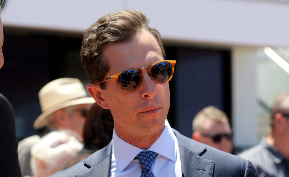 Man in a navy suit and tie wearing orange sunglasses at an outdoor event, with a crowd in the background.