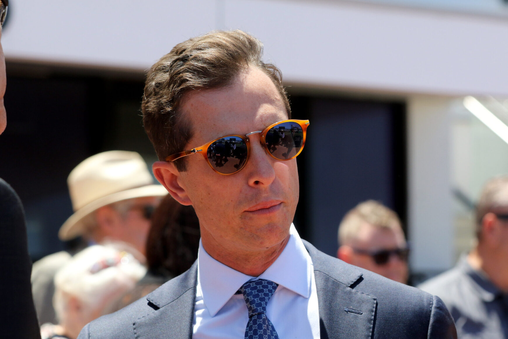 Man in a navy suit and tie wearing orange sunglasses at an outdoor event, with a crowd in the background.