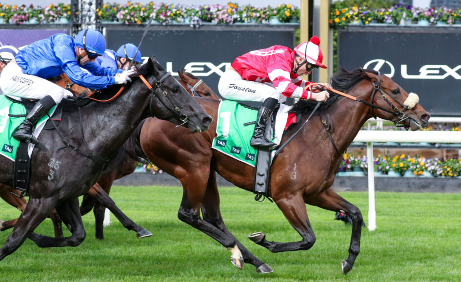 Two jockeys race side by side on thoroughbreds, one in red-and-white silks and the other in blue, on a green turf with sponsor boards behind them.