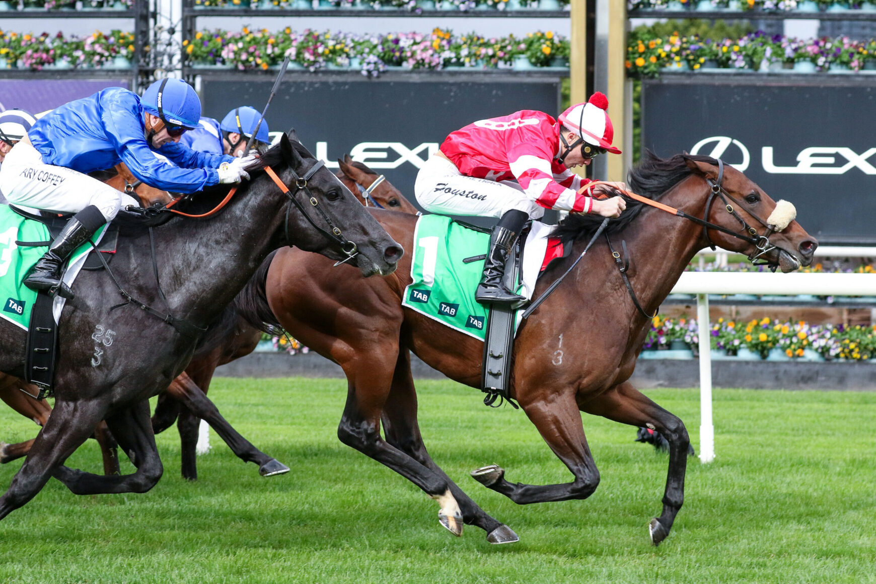 Two jockeys race side by side on thoroughbreds, one in red-and-white silks and the other in blue, on a green turf with sponsor boards behind them.