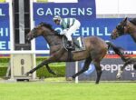 Bay racehorse with jockey in green-and-white silks, saddlecloth number 1, galloping on turf with blue signage behind him.