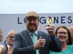 Man in a gray suit and flat cap lifts a gold trophy, smiling, as a cheering crowd applauds behind him at an award ceremony.