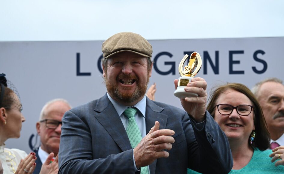 Man in a gray suit and flat cap lifts a gold trophy, smiling, as a cheering crowd applauds behind him at an award ceremony.