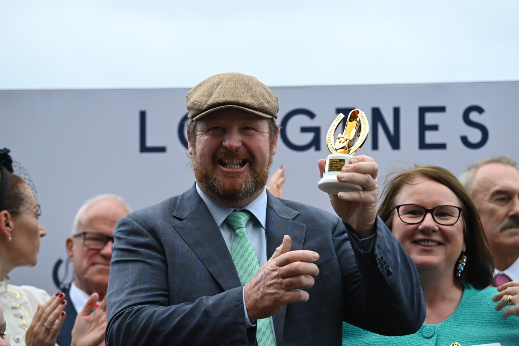Man in a gray suit and flat cap lifts a gold trophy, smiling, as a cheering crowd applauds behind him at an award ceremony.