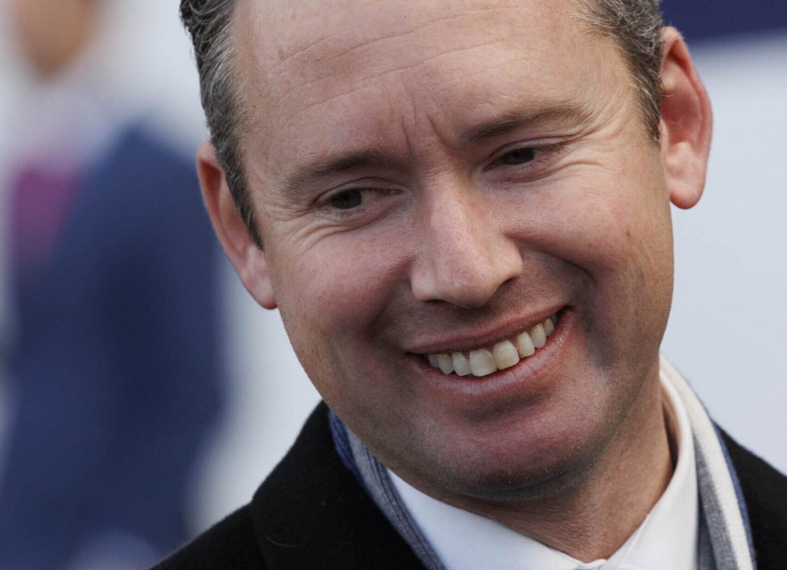 Close-up portrait of a smiling man with short dark hair, wearing a scarf and coat, looking slightly to the left.
