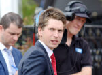 Man in a navy suit and red tie looks over his shoulder at an outdoor event, with a crew member in a headset blurred in the background.