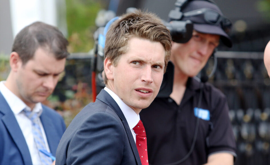 Man in a navy suit and red tie looks over his shoulder at an outdoor event, with a crew member in a headset blurred in the background.