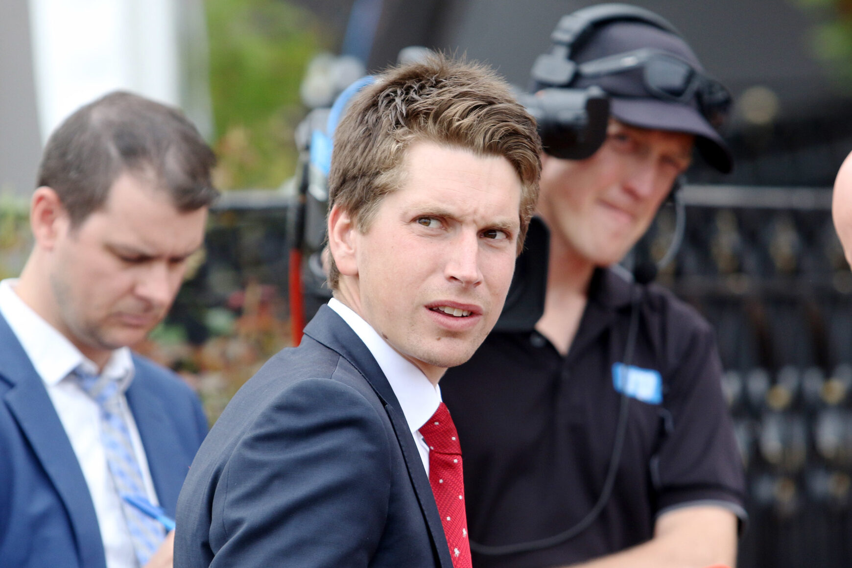 Man in a navy suit and red tie looks over his shoulder at an outdoor event, with a crew member in a headset blurred in the background.
