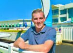 Man outdoors at an aquatic center, smiling and leaning on a white railing wearing a blue polo shirt.