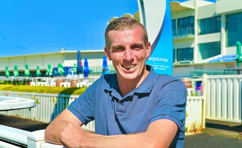 Man outdoors at an aquatic center, smiling and leaning on a white railing wearing a blue polo shirt.
