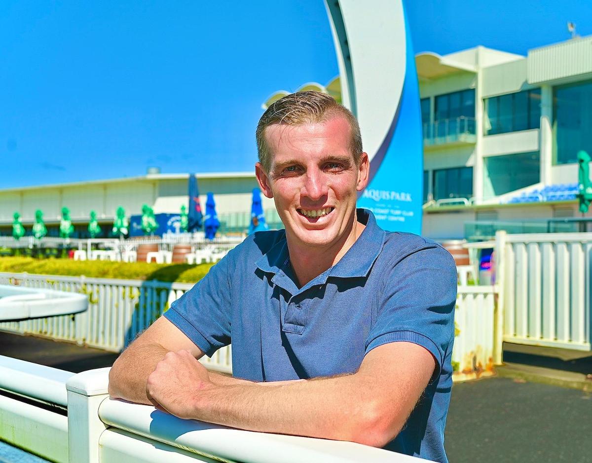 Man outdoors at an aquatic center, smiling and leaning on a white railing wearing a blue polo shirt.