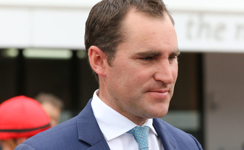Man in a navy suit with a light blue patterned tie, looking to the right at an outdoor event or venue.